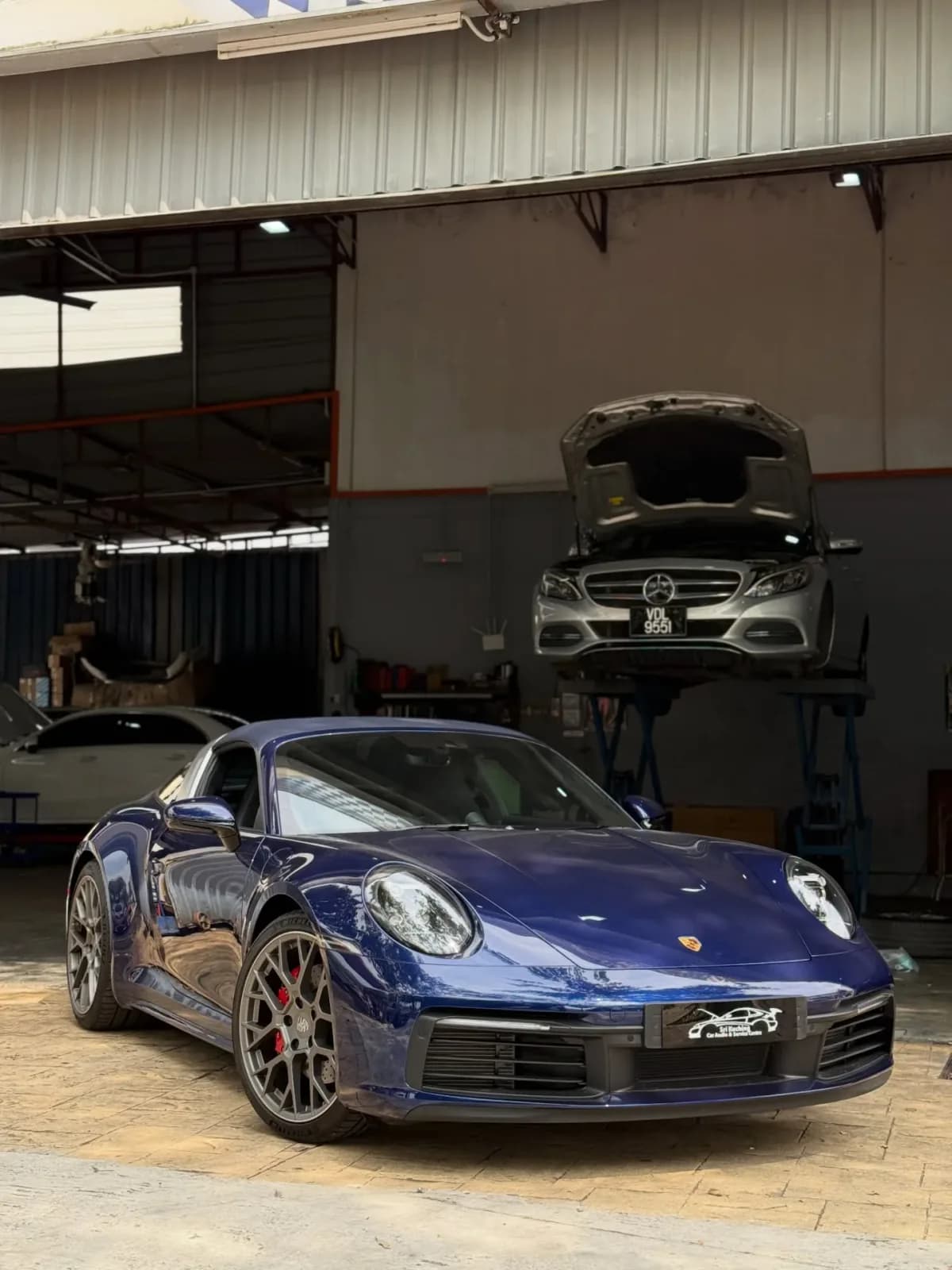 Porsche 911 at Sri Kuching workshop entrance with Mercedes on lift in background